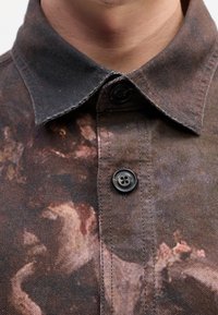 Gros plan sur le col et les boutons d'une chemise foncée avec un motif abstrait atténué aux tons marron doux et noir.