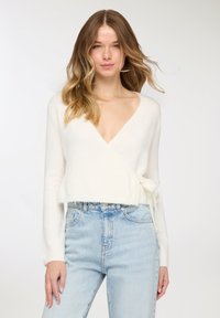 Maglione avvolgente bianco in tessuto morbido con maniche lunghe, scollo a V e dettaglio con laccio laterale, abbinato a jeans azzurri a vita alta.