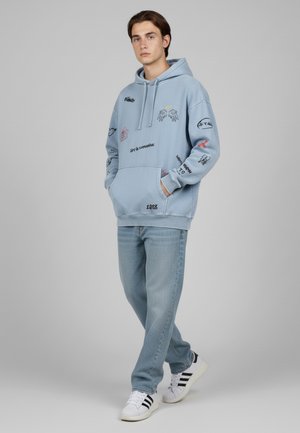 Light blue hoodie with various printed designs, front pocket, paired with light denim jeans and black-and-white sneakers.