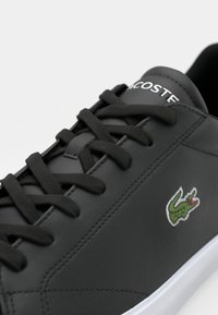 Basket en cuir noir avec lacets noirs, semelle blanche, écusson logo Lacoste sur le côté et inscription « LACOSTE » sur la languette.