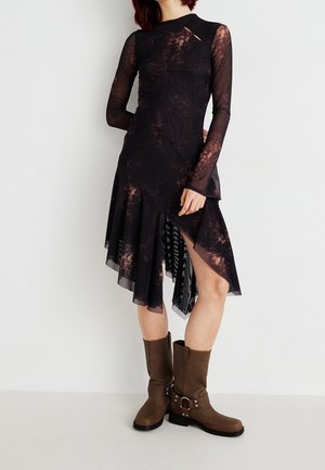 Femme portant une robe asymétrique tie-dye noire et marron avec des manches longues transparentes et des bottes marron mi-mollet à boucles.