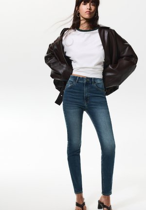 Skinny jeans in dark blue denim with contrast stitching, paired with a cropped white shirt and an oversized brown leather jacket.