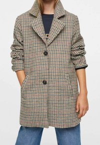 Ruudullinen takki, jossa on uurrettu kaulus, kolme nappia ja kaksi sivutaskua. Valmistettu tekstuurillisesta kankaasta, jossa on beigen, punaisen ja mustan sävyjä.