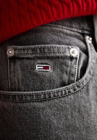 Jeans de mezclilla gris con un bolsillo frontal que presenta un pequeño logo bordado en rojo, blanco y azul, junto con acentos de remaches metálicos.