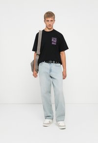 Young man wearing a black graphic T-shirt, light blue jeans, white sneakers, carrying a gray shoulder bag, standing against a white wall.
