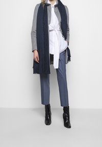 Manteau gris avec des boutons, écharpe marine, chemise blanche avec un nœud, et pantalon bleu à motifs. Bottines noires avec une texture finition.