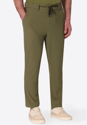 Uomo con pantaloni a coste verde oliva con vita con bottone e coulisse, sneakers beige, in piedi con le mani nelle tasche.