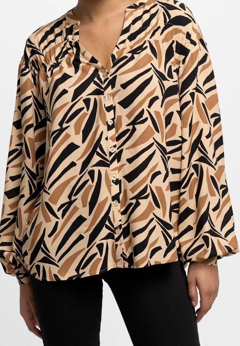 Femme portant un chemisier beige avec des motifs de feuilles abstraits noirs et marron et des boutons, associé à un pantalon noir, vue du cou aux hanches.