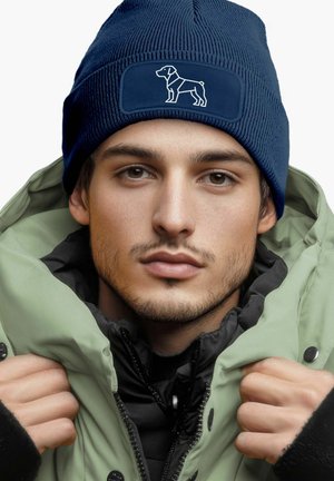 Navy knitted beanie with a rectangular patch featuring a white dog silhouette. Model wears a green puffer jacket layered over a black coat.