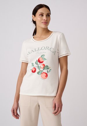 Femme portant un t-shirt blanc avec un imprimé de fruits rouges et de feuilles vertes et le texte "Mallorca España", associé à un pantalon beige.