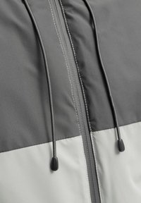Grau- und kremfarbene Jacke mit einem Vorderreißverschluss, ausgestattet mit verstellbaren Zugbändern, einer strukturierten Oberfläche und einem Kontrast-Farbblockdesign.