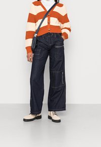 Czerwono-kremowy sweter w paski z guzikami, zestawiony z ciemnymi, szerokimi spodniami cargo z denimu z wieloma kieszeniami i zamkami błyskawicznymi. Beżowe botki.