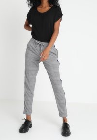 Haut noir à manches courtes, pantalon à carreaux gris avec des bandes latérales bleu marine. Le pantalon a une taille élastique et des chevilles resserrées. Chaussures noires.