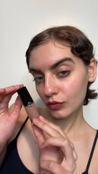 Naakte vloeibare lipgloss in een doorzichtige cilindrische tube met een zwarte dop en een zilveren accenten, vastgehouden door een persoon met een bleke huid.