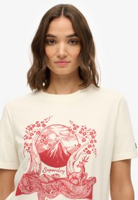 T-shirt crème avec un design graphique rouge représentant une montagne, un soleil, un cerf et des éléments floraux, accompagné du texte "Superdry". Manches courtes et coupe décontractée.