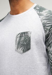 Uomo che indossa una camicia grigia chiaro con maniche a motivo foglie di palma verde scuro e taschino sul petto, mostrato dalla gola fino a metà tronco.