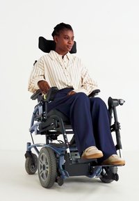 Fauteuil roulant électrique avec un cadre bleu, un siège noir et de grandes roues arrière. L'utilisateur porte une chemise rayée et un pantalon foncé, avec des chaussures beiges.