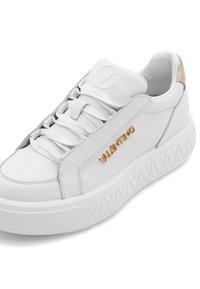 Sneaker in pelle bianca con suola chunky, caratterizzato da un logo dorato "VALENTINO", lacci tono su tono e un accento dorato metallico sul tallone.