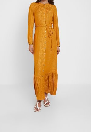 Femme portant une robe longue à manches longues, boutonnée, de couleur jaune moutarde avec une ceinture à la taille et un imprimé léopard subtil, assortie à des sandales blanches à brides.