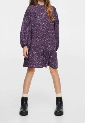 Niña con un vestido largo de manga larga morado con flores, calcetines color crema y botas negras hasta el tobillo, de pie con las piernas separadas.