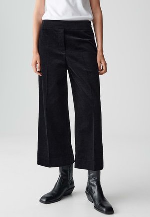 Persoon in zwarte wijde corduroy broek, zwarte enkellaarsjes en een witte ingestoken top, staand op een effen lichte achtergrond.
