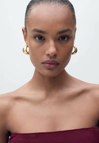 Model met een egale huid, gekleed in een bordeaux strapless top, gouden hoepel oorbellen, en stijlvol haar dat naar achteren is vastgebonden, tegen een neutrale achtergrond.