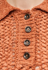 Κοντινό πλάνο πορτοκαλί πλεκτού ενδύματος με υφή ανοιχτής ύφανσης και τέσσερα σκούρα καφέ κουμπιά στο κέντρο μπροστά.