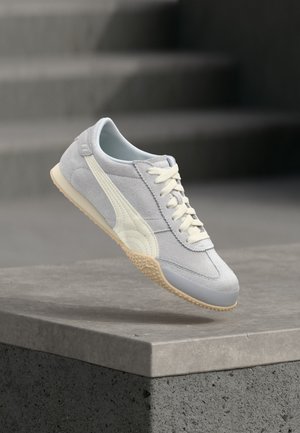 Pantof sport din piele întoarsă gri deschis cu dungi albe din piele și talpă de gumă, suspendat deasupra unui prag din beton cu trepte gri neclare în fundal.