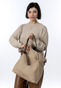 Sac à main beige à texture douce avec des anses en cuir marron et un accent à pompon. Design structuré avec une forme effilée et une fermeture éclair.