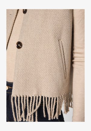 Gilet in maglia beige con texture, chiusura con bottoni, tasca laterale e orlo con frange, indossato sopra una maglietta a maniche lunghe crema e jeans in denim scuro.