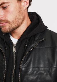 Černá kožená bunda s matným povrchem, kterou zdobí zip na přední straně a dvě viditelné kapsy, vrstvená přes černou mikinu.