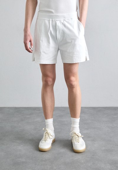 White cotton shorts with a soft texture, elastic waist, side pockets, and slight side slits. Paired with white sneakers and socks.