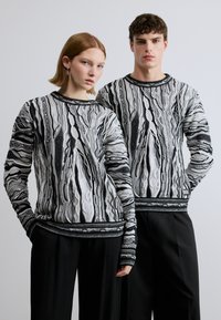 Pulls tricotés à motifs en noir et blanc avec manches longues, présentant des designs texturés et des finitions noires contrastantes, assortis à des pantalons décontractés.