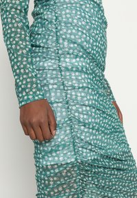 Vestido verde-água transparente com padrão floral branco, apresentando pregas ao longo das laterais e mangas longas, destacando um acabamento texturizado.