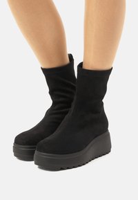 Botas de tobillo de ante negro con una textura suave, forma cónica y una suela de plataforma gruesa y contrastante que presenta un patrón en zigzag.