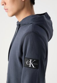 Tamsiai mėlyna bliuzono su tekstūriniu paviršiumi, kuriame yra juodas stačiakampis lopas ant rankovės su baltu "CK" logotipu ir prekės ženklu.
