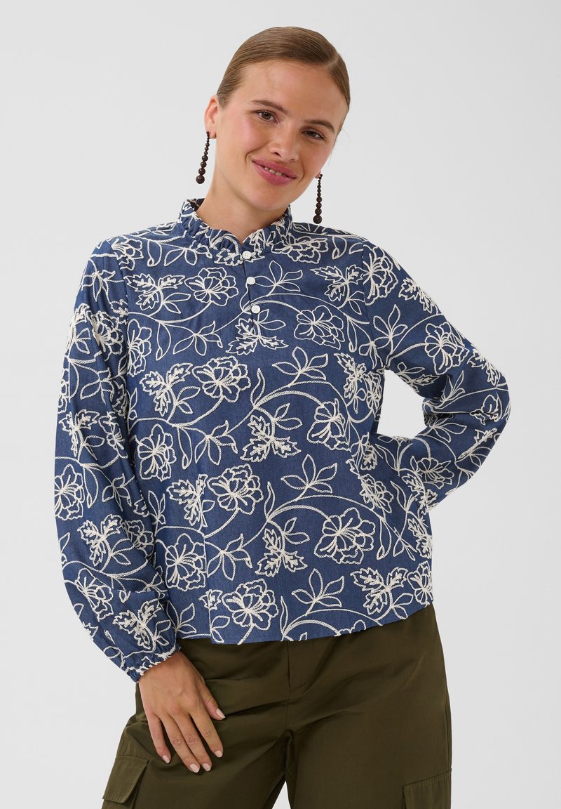 Femme portant un chemisier bleu avec une broderie florale blanche et un pantalon vert olive, souriante, une main sur la hanche et l'autre reposant sur la cuisse.