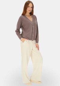 Jasnobrązowy sweter z wełny z dekoltem w serek, z guzikami z przodu oraz ściągaczami przy mankietach. W połączeniu z jasnobeżowymi spodniami z szerokimi nogawkami.