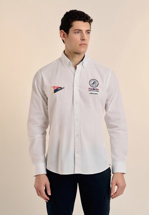 Camisa de botones blanca de manga larga con parches de logo; presenta un collar clásico, un dobladillo recto y una textura de tela suave de algodón.