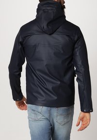 Marinblå vattentät jacka med huva, med en slät textur och strömlinjeformat design. Bakifrån syns axelsömmar och justerbara manschetter.