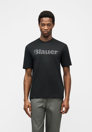 Jeune homme portant un t-shirt noir avec le logo « Blauer » et un pantalon gris, se tenant face à l'avant sur un fond blanc uni.