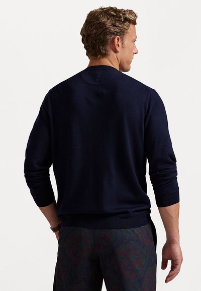 Bărbat cu păr creț, îmbrăcat cu un pulover lung cu mâneci navy și pantaloni în model paisley închise la culoare, întors cu spatele pe un fundal alb simplu.