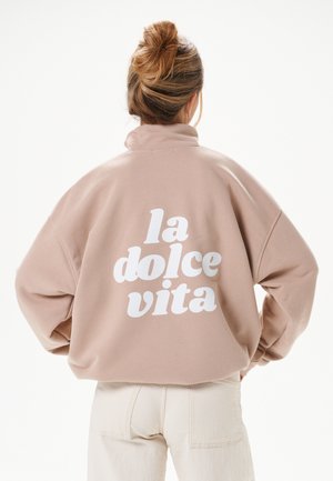 Vrouw met blond haar in een knot draagt een beige oversized jas met op de achterkant in grote witte letters "la dolce vita" gedrukt.