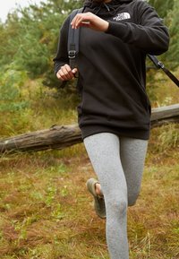 Svart huvtröja med logotyp, grå leggings och gröna sneakers. Springer i en utomhusmiljö med gräs, buskar och en fallen stock i bakgrunden.