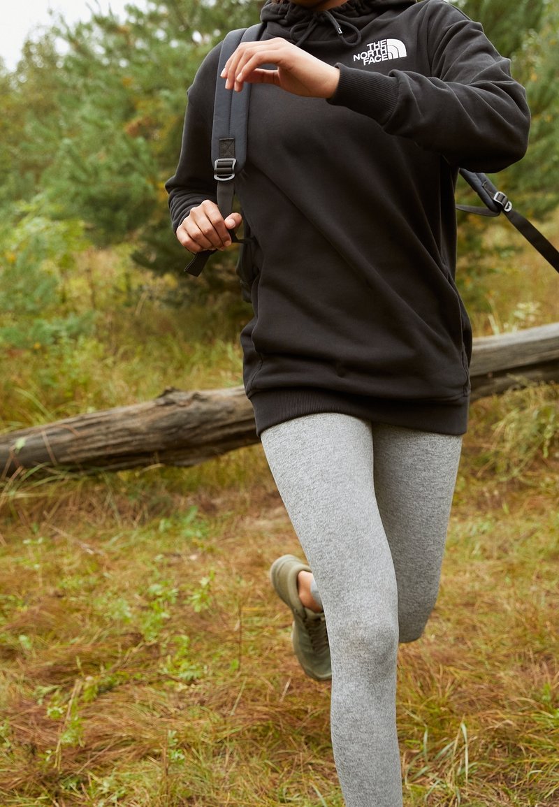 Svart huvtröja med logotyp, grå leggings och gröna sneakers. Springer i en utomhusmiljö med gräs, buskar och en fallen stock i bakgrunden.