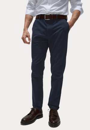 Homme portant un pantalon bleu marine, une ceinture en cuir marron, des chaussures marron foncé à lacets, et une chemise bleu clair à boutons avec les manches retroussées.