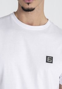 Biely bavlnený tričko s okrúhlym výstrihom a malým čiernym štvorcovým logom so znakmi "K" a "G" na ľavej strane hrudi.