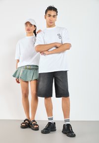 Dos modelos están de pie uno al lado del otro. Uno lleva una camiseta blanca de gran tamaño y una falda de mezclilla. El otro, con una camiseta blanca y pantalones cortos negros, lleva zapatillas.