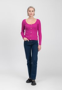 Fuchsia geribbelde trui met lange mouwen en een ronde halslijn, voorzien van gouden knopen aan de voorkant, gecombineerd met donkere spijkerbroeken en zwarte chunky schoenen.