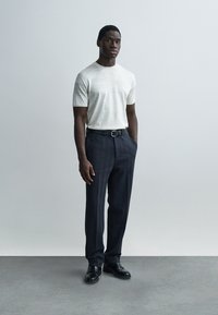 Ανοιχτό γκρίζο κοντομάνικο πλεκτό πουκάμισο με λεπτές ρίγες, συνδυασμένο με σκούρο παντελόνι με μοτίβα και μαύρα δερμάτινα παπούτσια, στέκεται μπροστά σε απλό φόντο.